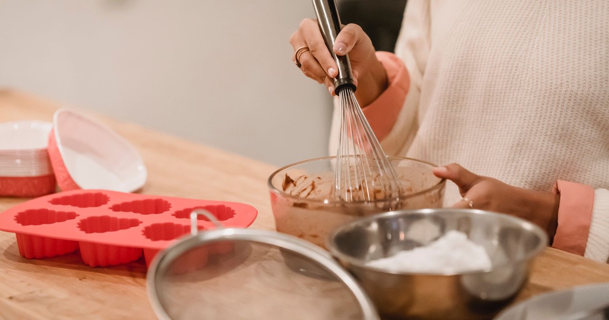 Unrecognizable female baker with whisk mixing ingredients for pastry while standing at table with molds and flour during cooking in kitchen