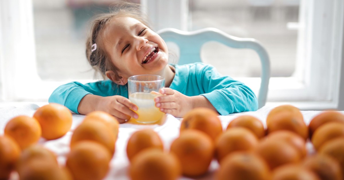 A cheerful child smiling while holding a glass of fresh orange juice surrounded by oranges indoors.