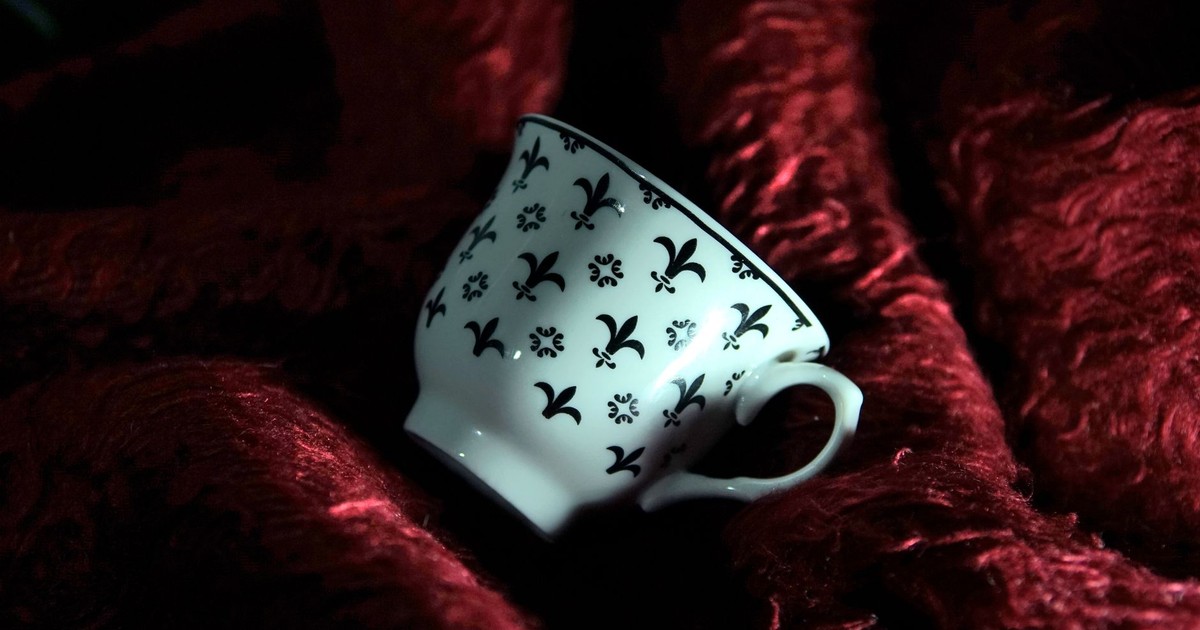 A white teacup with fleur-de-lis pattern resting on rich red fabric, creating a luxurious vibe.