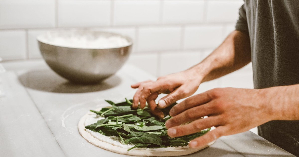 A chef adds fresh spinach to a pizza dough in a kitchen setting.