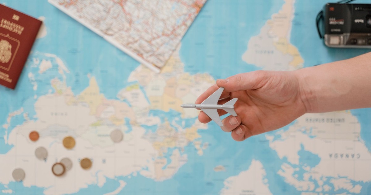 Hand holding a toy airplane over a world map, surrounded by coins, camera, and passport.