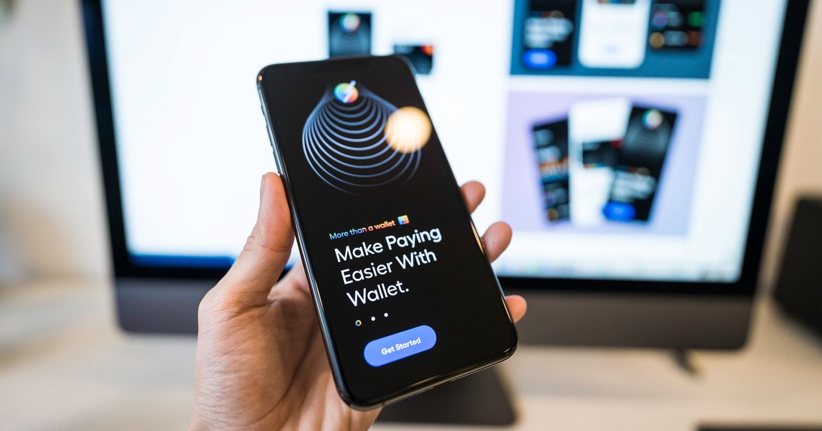 Hand holding smartphone displaying digital wallet app interface, blurred monitor in background.