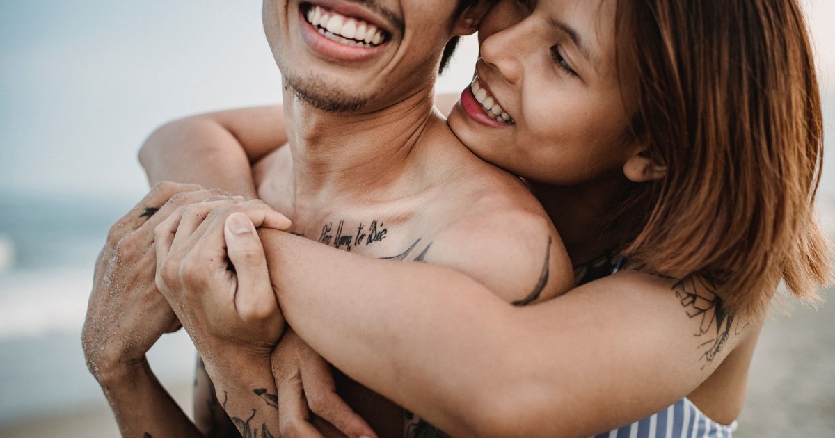 A young couple embraces playfully on the beach, showcasing tattoos and joyful smiles.