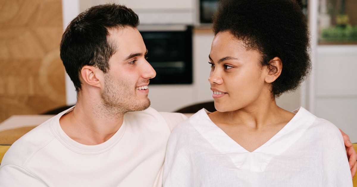 A young interracial couple sitting close together inside a cozy home setting, smiling warmly at each other.