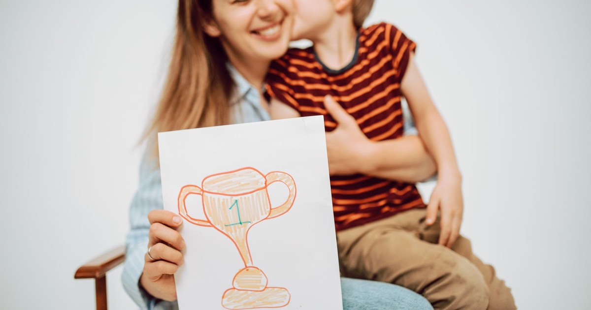A cheerful mother receives a hug and a drawing labeled 'Best Mom' from her young son.