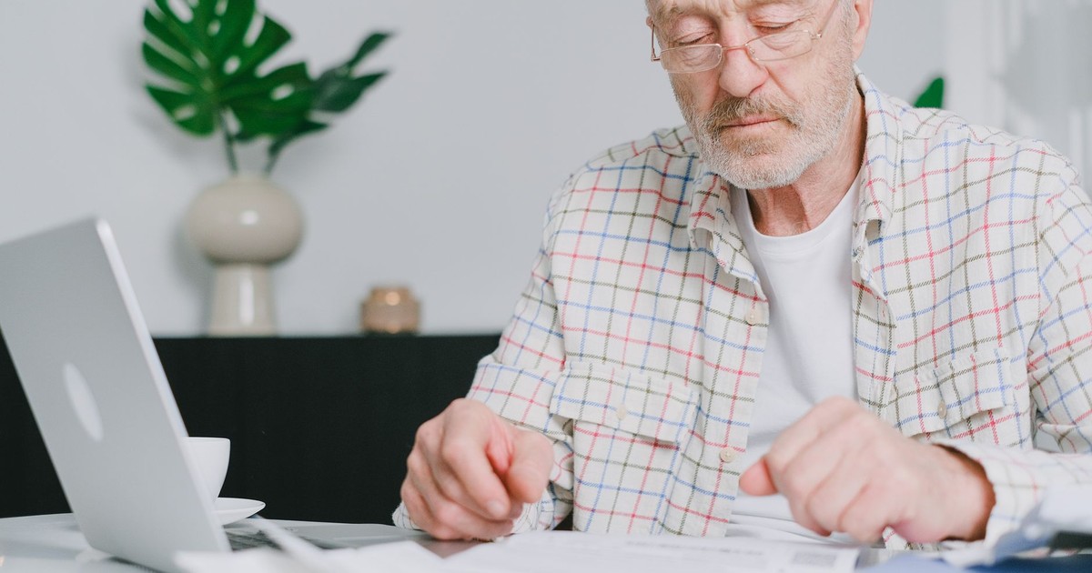 Elderly man with eyeglasses focused on paperwork at home, using laptop.