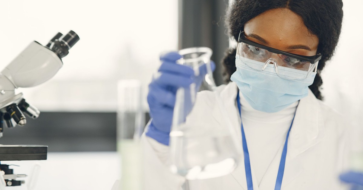 Woman scientist in lab coat and mask conducting experiment with microscope and beaker.