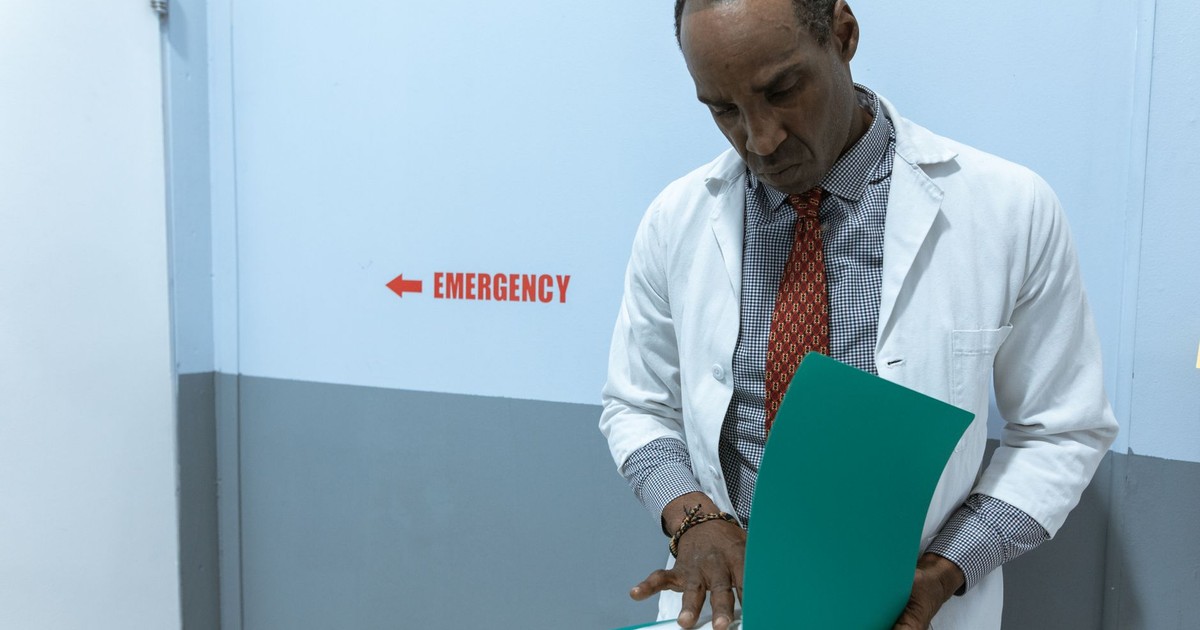 A doctor in a lab coat examines documents near the emergency room sign.