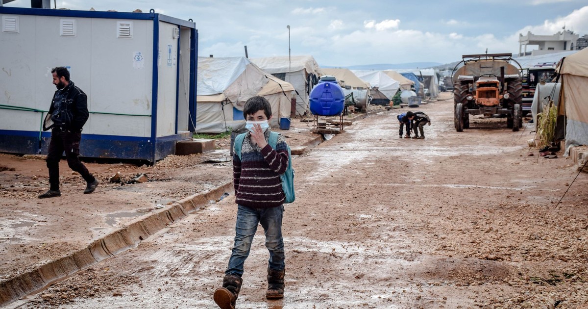 Full body of ethnic boy in protective mask strolling on dirty path along shabby tents in poor district of countryside during pandemic