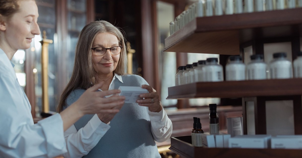 Elderly woman receiving assistance from a pharmacist in a pharmacy with product shelves.
