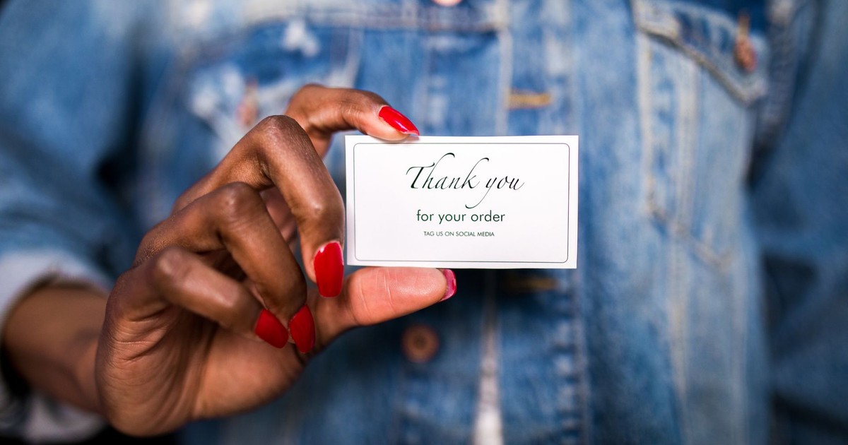 A woman in a denim jacket holds a 'Thank You' card with red nails.