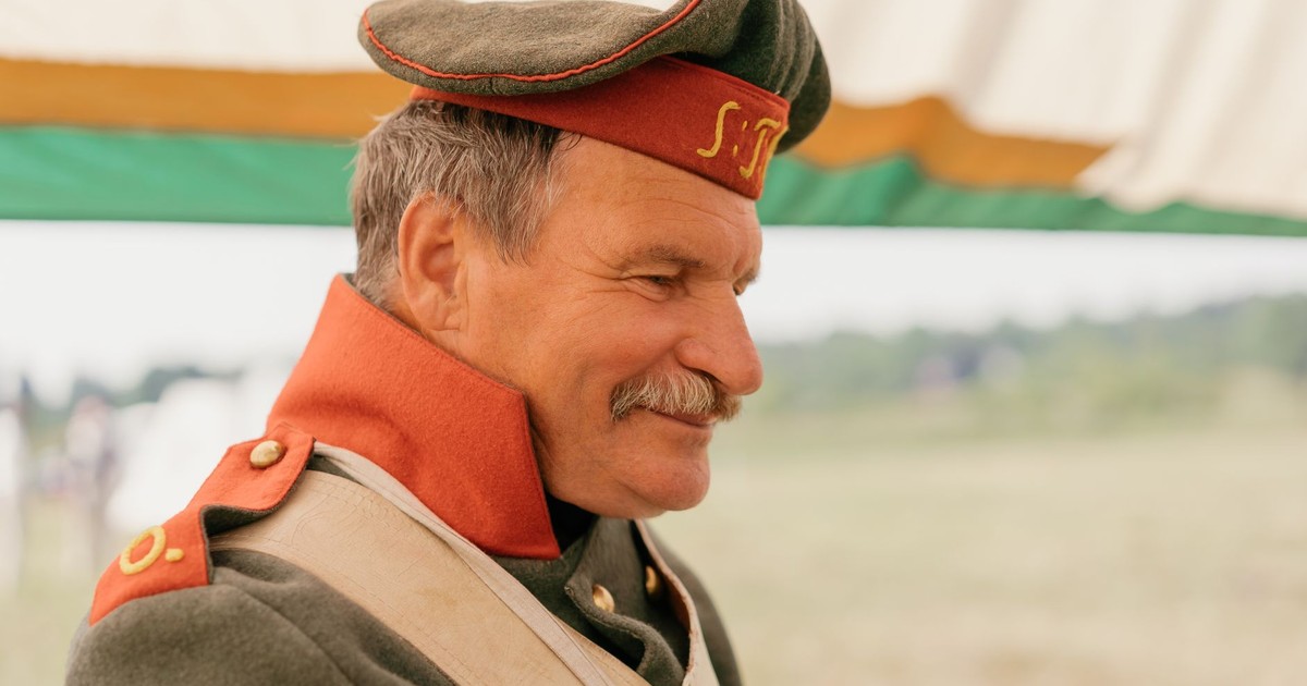 Elderly man in vintage military uniform smiling outdoors at a reenactment event.
