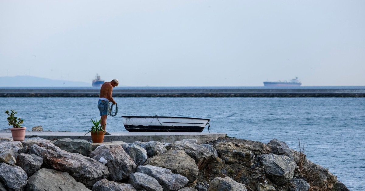 A man prepares a boat on a rocky shoreline with cargo ships in the background.