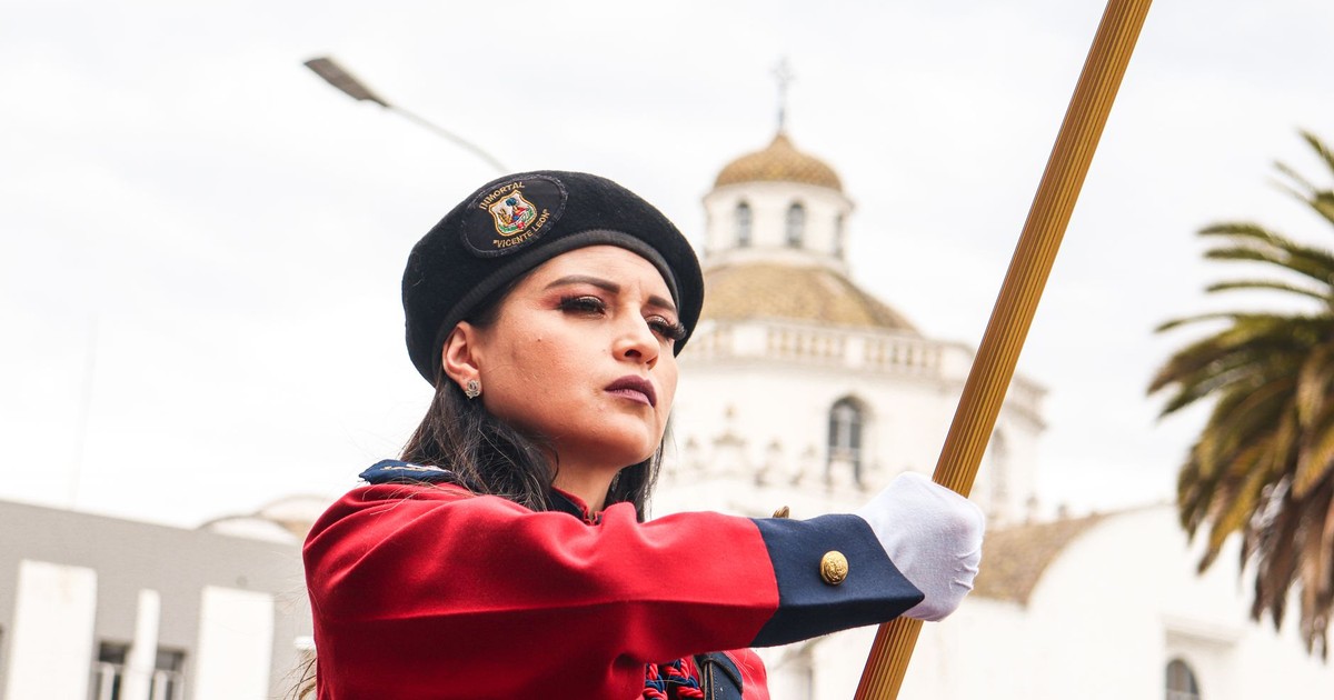 Young woman in military-style uniform holding a flag during a parade in Ecuador, with a historic building in the background.