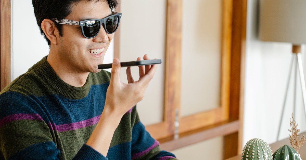 Young man wearing sunglasses smiles while using voice features on his smartphone indoors.