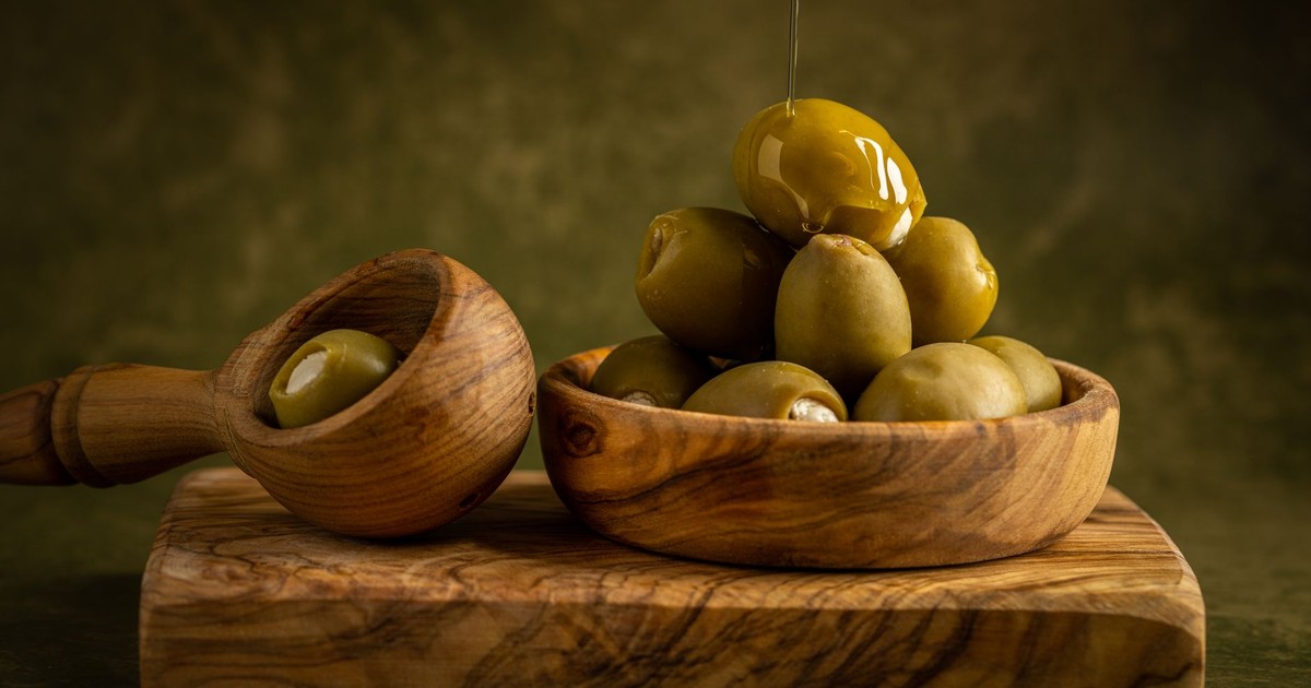 An artistic composition of green olives drizzled with oil in a wooden bowl on a rustic cutting board.