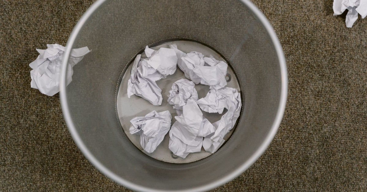 Overhead view of a trash can filled with crumpled paper on a carpeted floor.