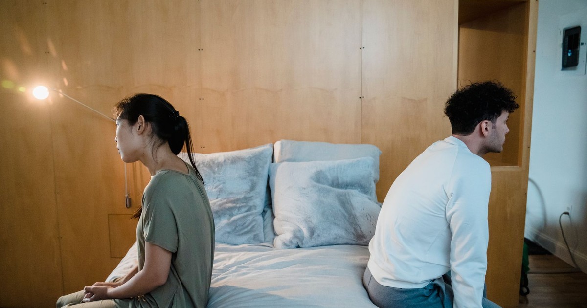 A couple sits back to back on a bed, showing tension and misunderstanding in a cozy bedroom.