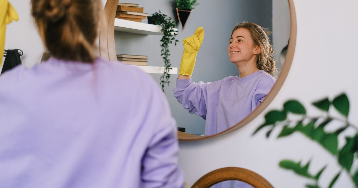 Smiling woman cleaning mirror with yellow gloves indoors, reflecting her image.