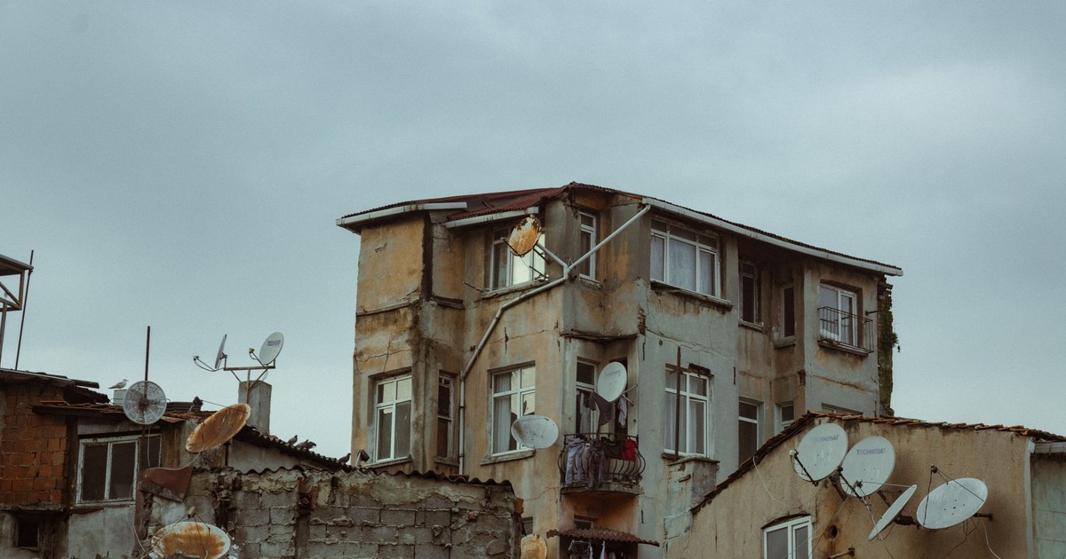 A worn-down residential building with numerous satellite dishes under a cloudy sky.