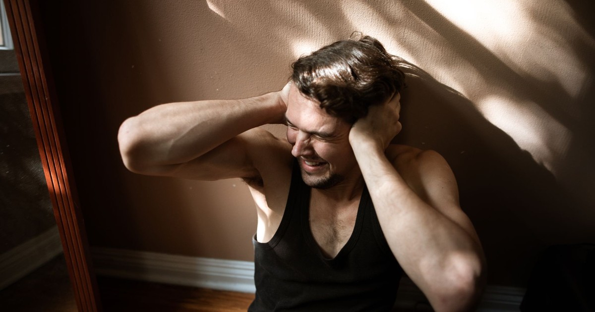 A man sitting on the floor indoors, showing signs of stress by covering his ears.