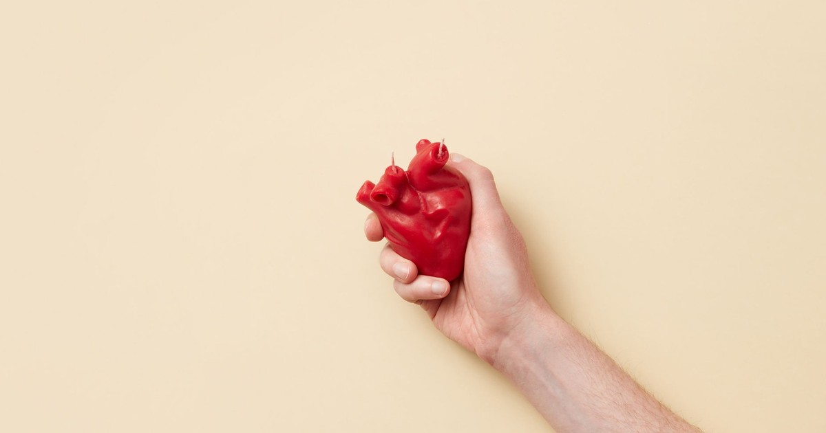 A realistic red anatomical heart model held firmly in a hand against a beige background.
