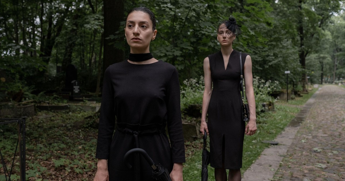 Two women dressed in black stand in a serene cemetery pathway amidst lush greenery.