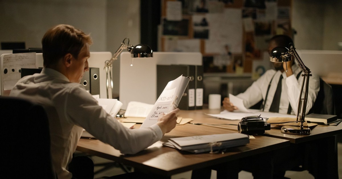 Two detectives analyzing documents in an office setting under dim lighting, focusing on investigation.