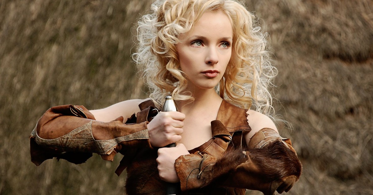 A blonde woman in warrior attire holding a sword, set against a rustic outdoor background in Kyiv.