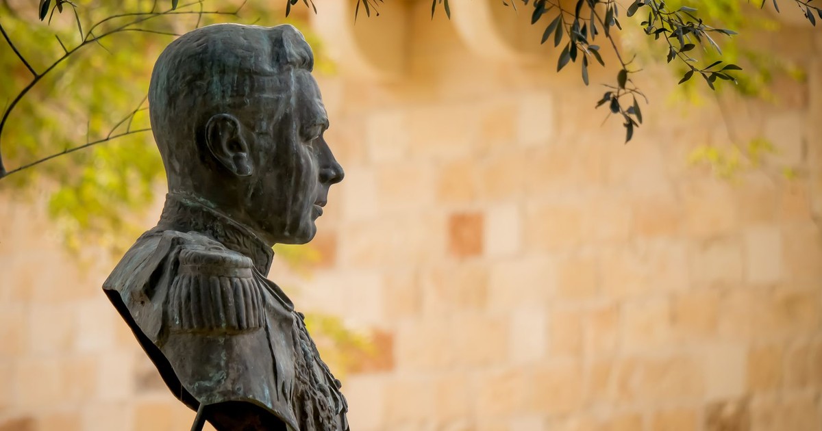 A classical bronze bust in profile view outdoors, bathed in warm sunlight, surrounded by greenery.