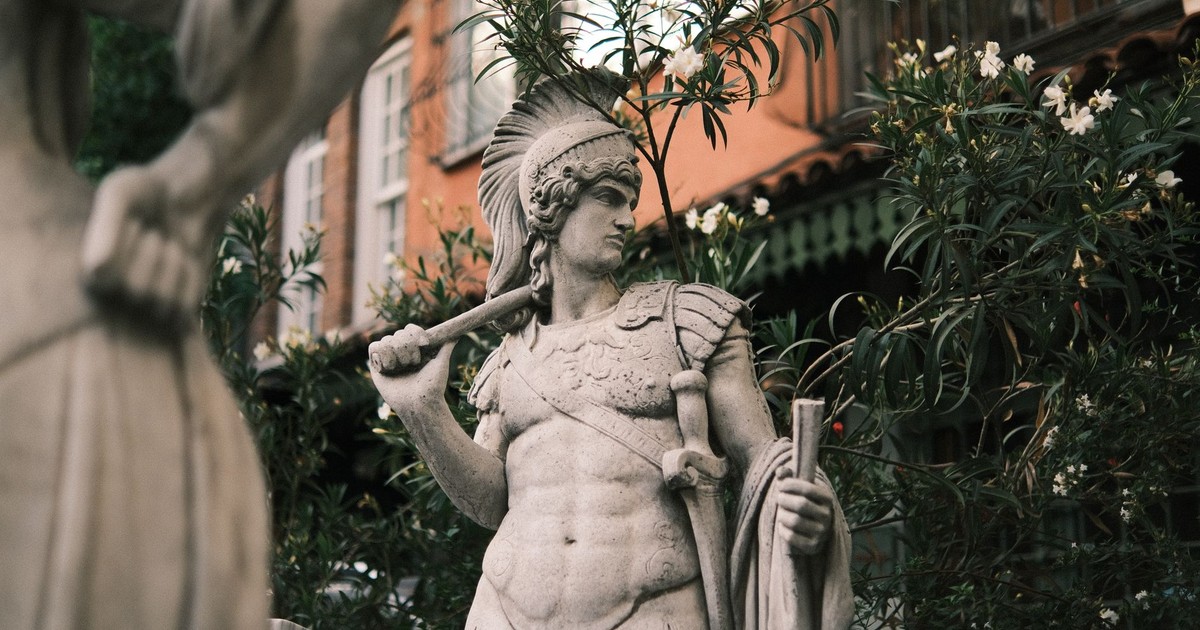 Stone statue of ancient warrior set against urban backdrop with foliage.
