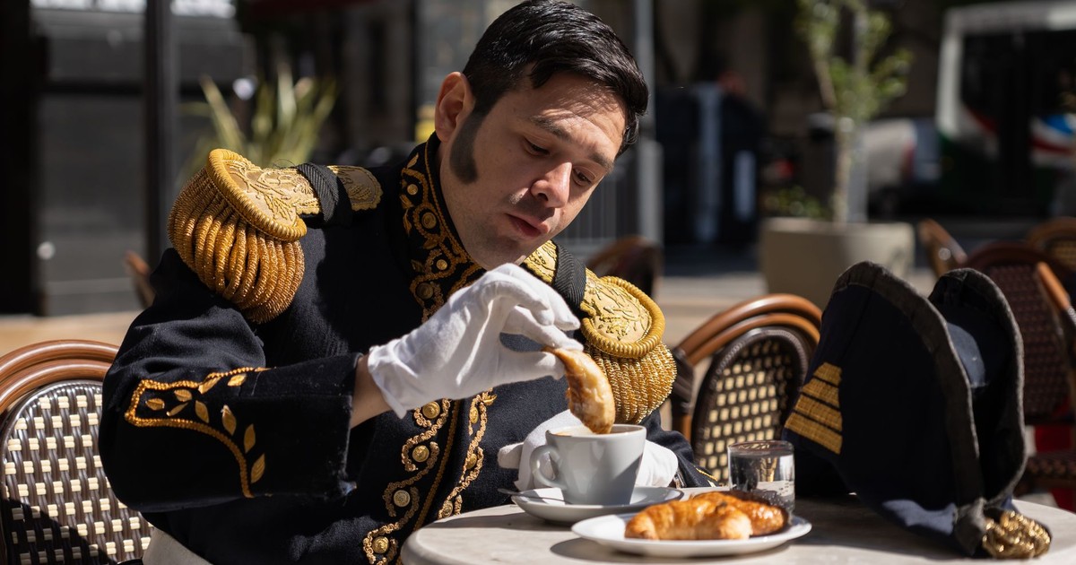 A man in a historical military uniform enjoys coffee and a croissant at an outdoor cafe in bright daylight.