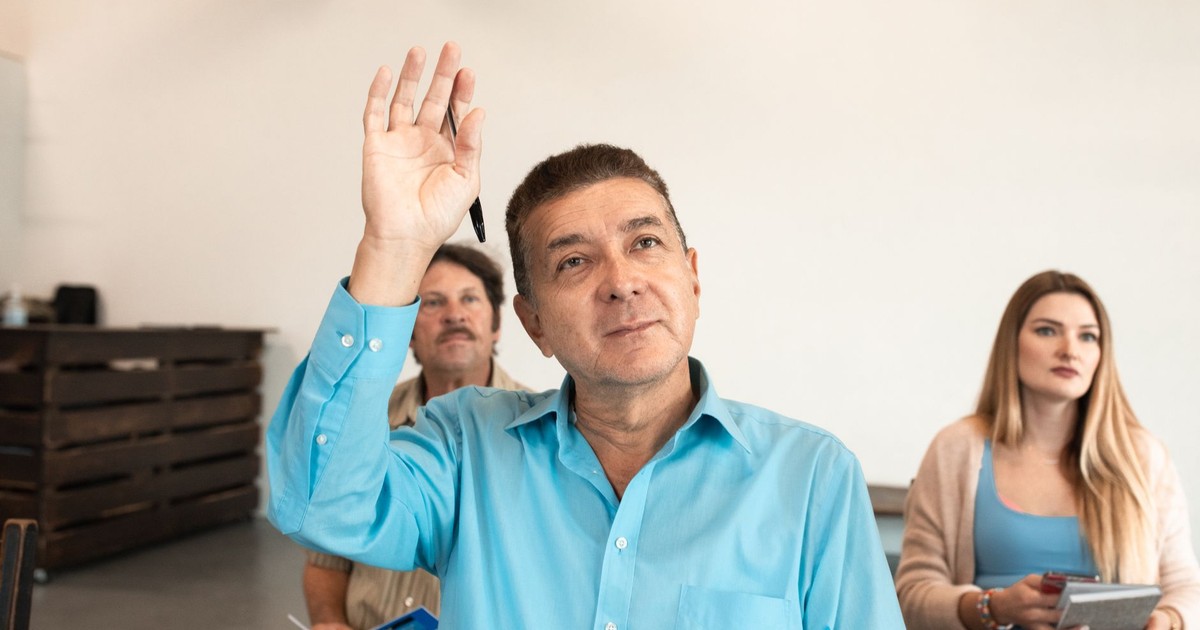 Adults learning in a classroom, with a man raising his hand to pose a question.
