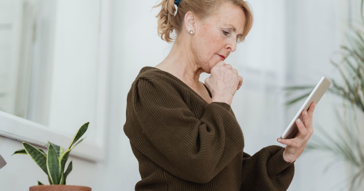 Side view of thoughtful elderly female in sweater with cellphone touching chin against potted plant in house room