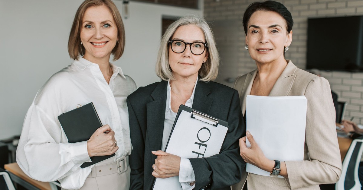 Three confident businesswomen in professional attire holding papers in an office setting.