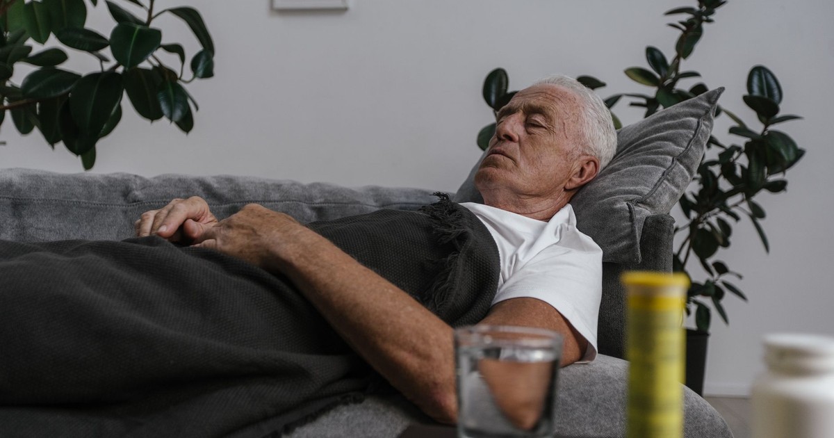An elderly man sleeping on a couch with a blanket and glass of water nearby.