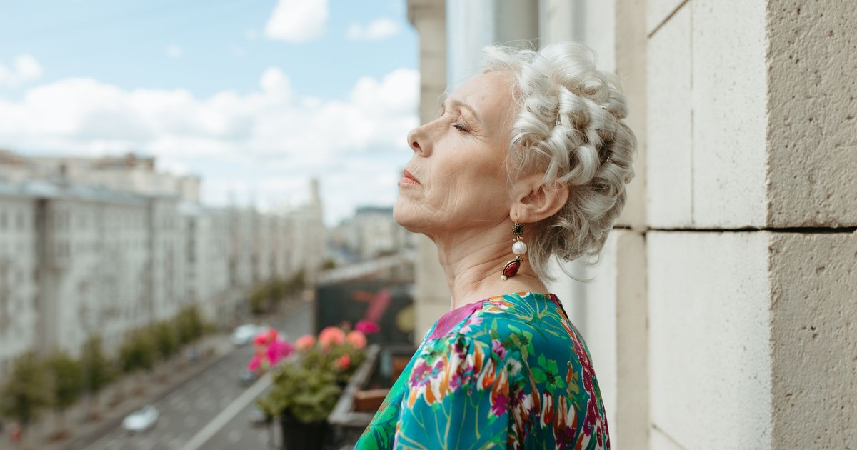 Senior woman with white hair and earrings enjoys a view from a balcony in the city.