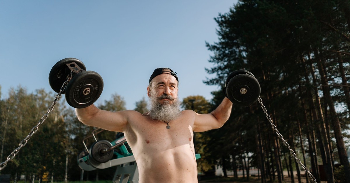 A senior man with a beard exercises shirtless outdoors with dumbbells, emphasizing fitness and determination.
