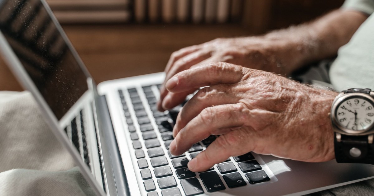 Close-up of a senior adult's hands typing on a laptop, wearing a wristwatch, in a home office setup.