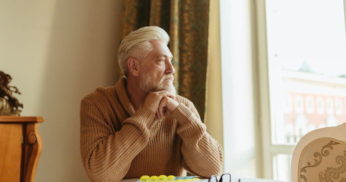 Elderly man with a beard sitting by a window, looking thoughtful.