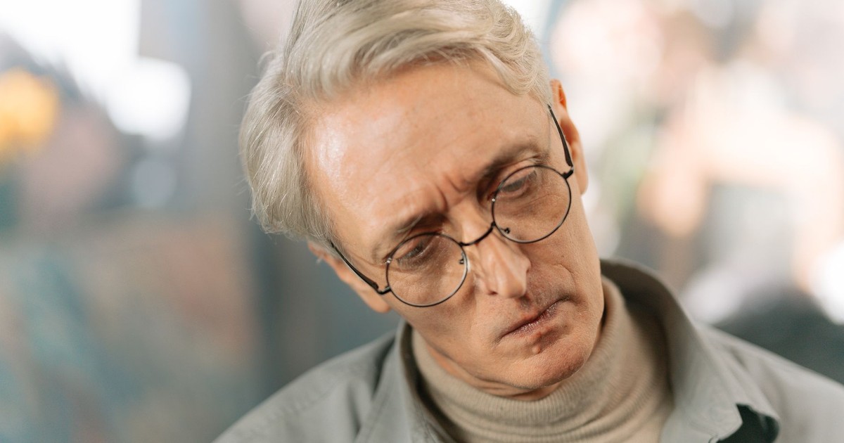 Elderly man with glasses and gray hair appearing pensive and thoughtful indoors.