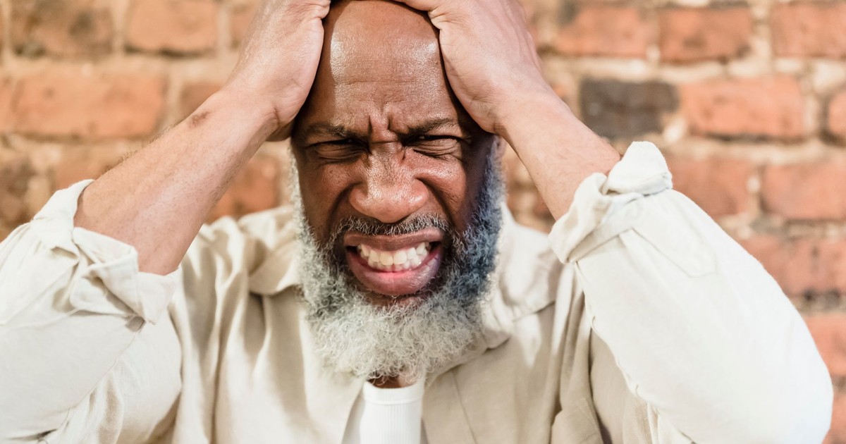 Close-up portrait of an adult man expressing pain and stress, holding head against a brick wall.