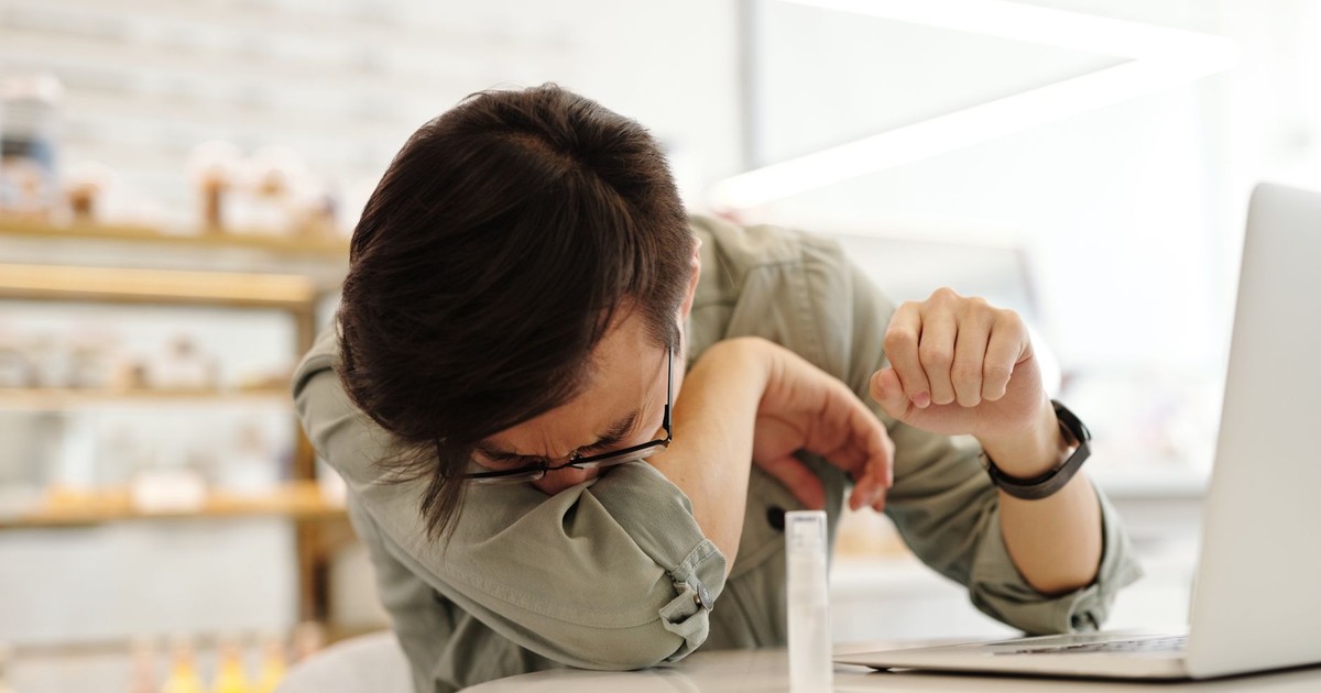 An adult man sneezes into his arm near a laptop in a cafe, illustrating illness prevention.