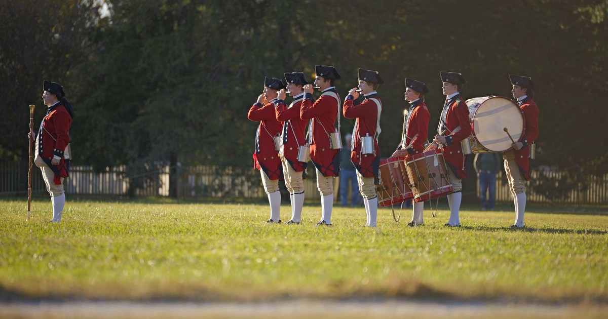 Colonial-era military band reenacting history in open field.