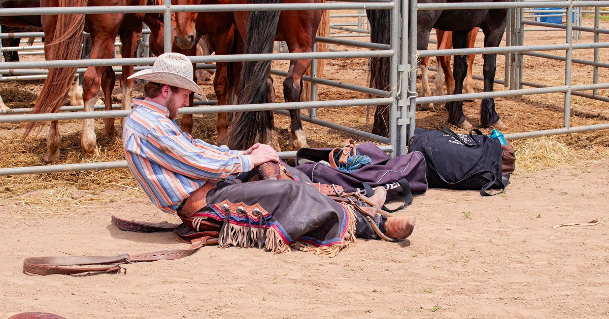 A cowboy in traditional attire resting beside a group of horses in a corral.