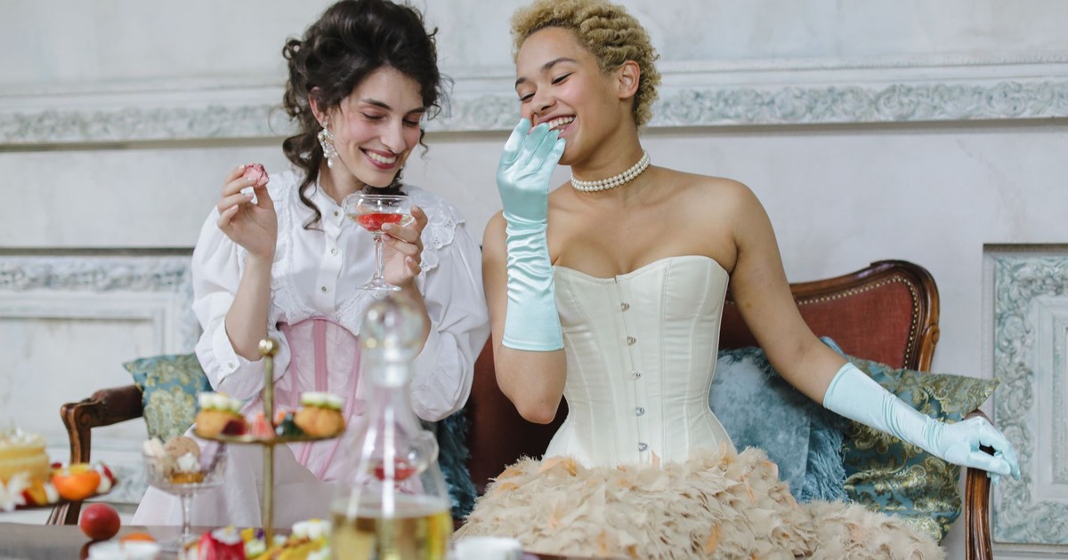 Two women in corsets enjoy an elegant afternoon tea setting indoors.