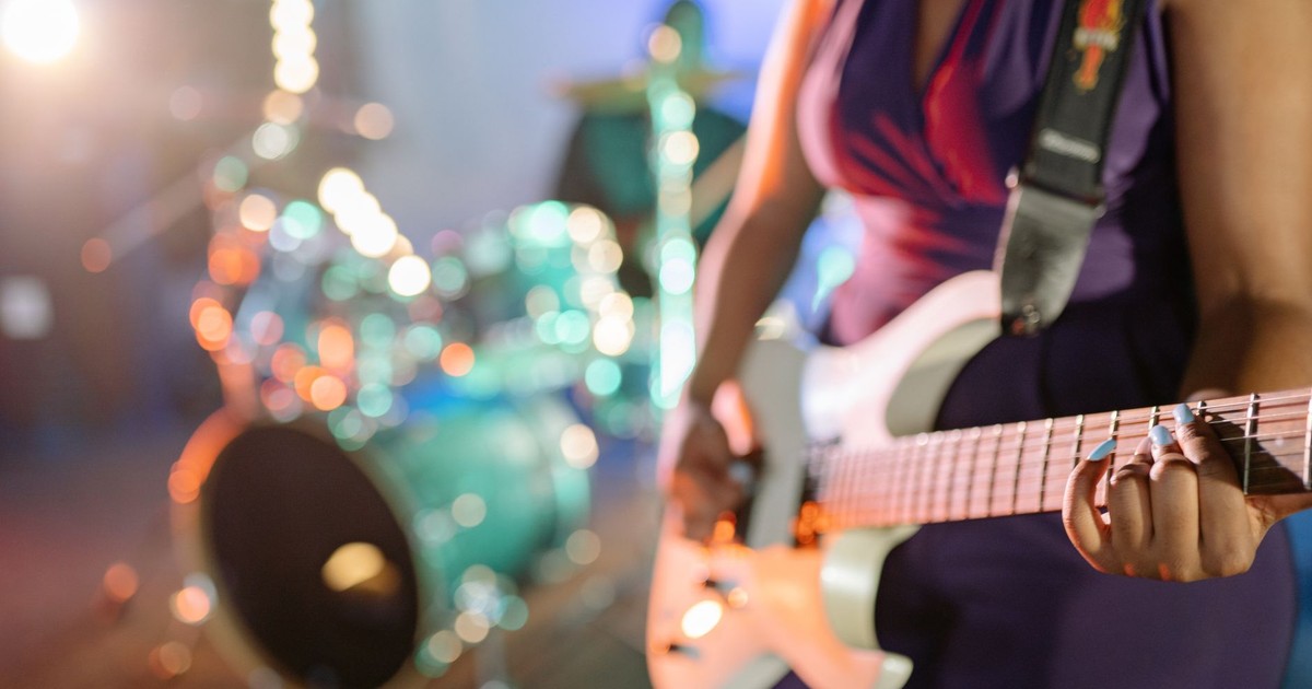 Close-up of a female guitarist playing on stage with drum set in background.
