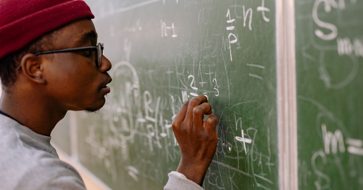 A young man with eyeglasses and a red beanie solving math problems on a chalkboard.