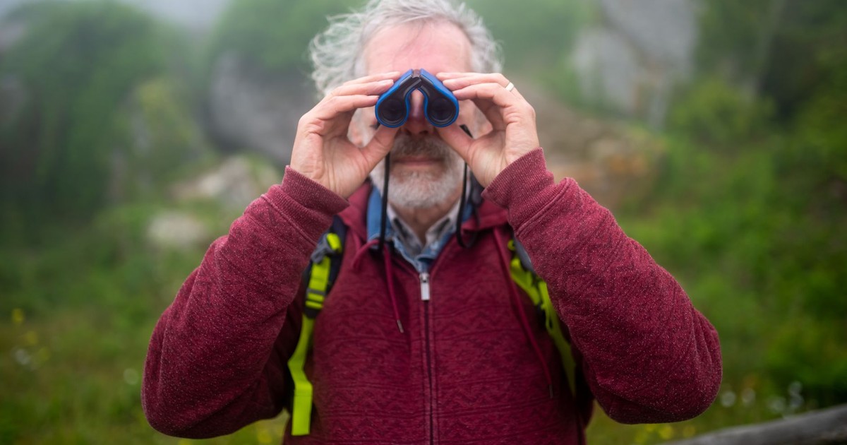 Elderly man hiking in lush green Portugal, observing nature with binoculars.