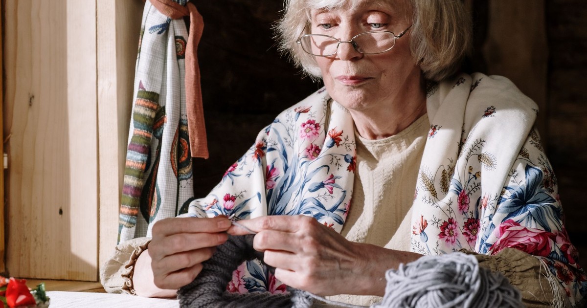 Senior woman knitting with gray yarn near a sunlit window, showcasing peaceful retirement activities.
